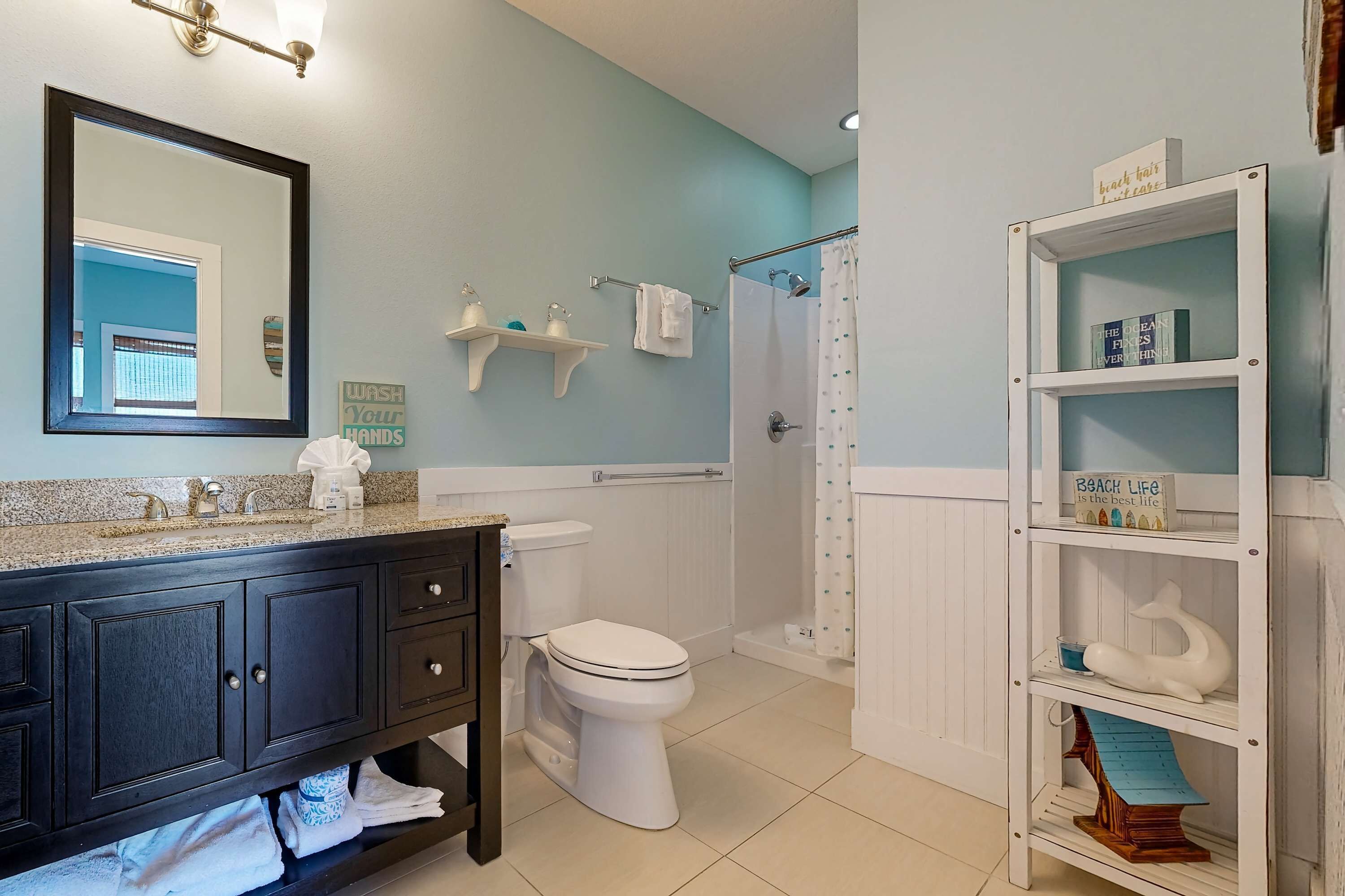 Un bagno con un mobile vanitario in legno scuro, un water bianco, una doccia con tenda bianca e uno scaffale bianco decorato.