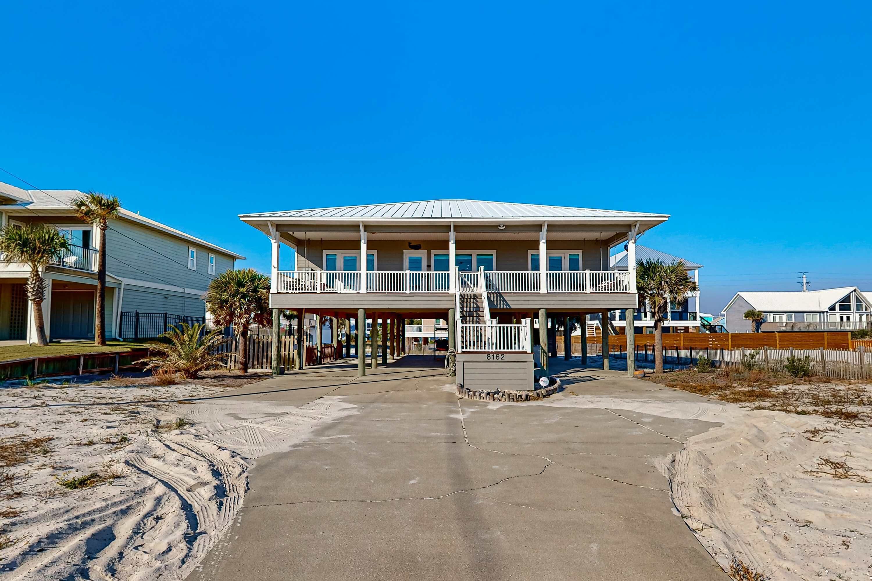 A beachfront house with a balcony, a covered porch, and a sandy driveway.