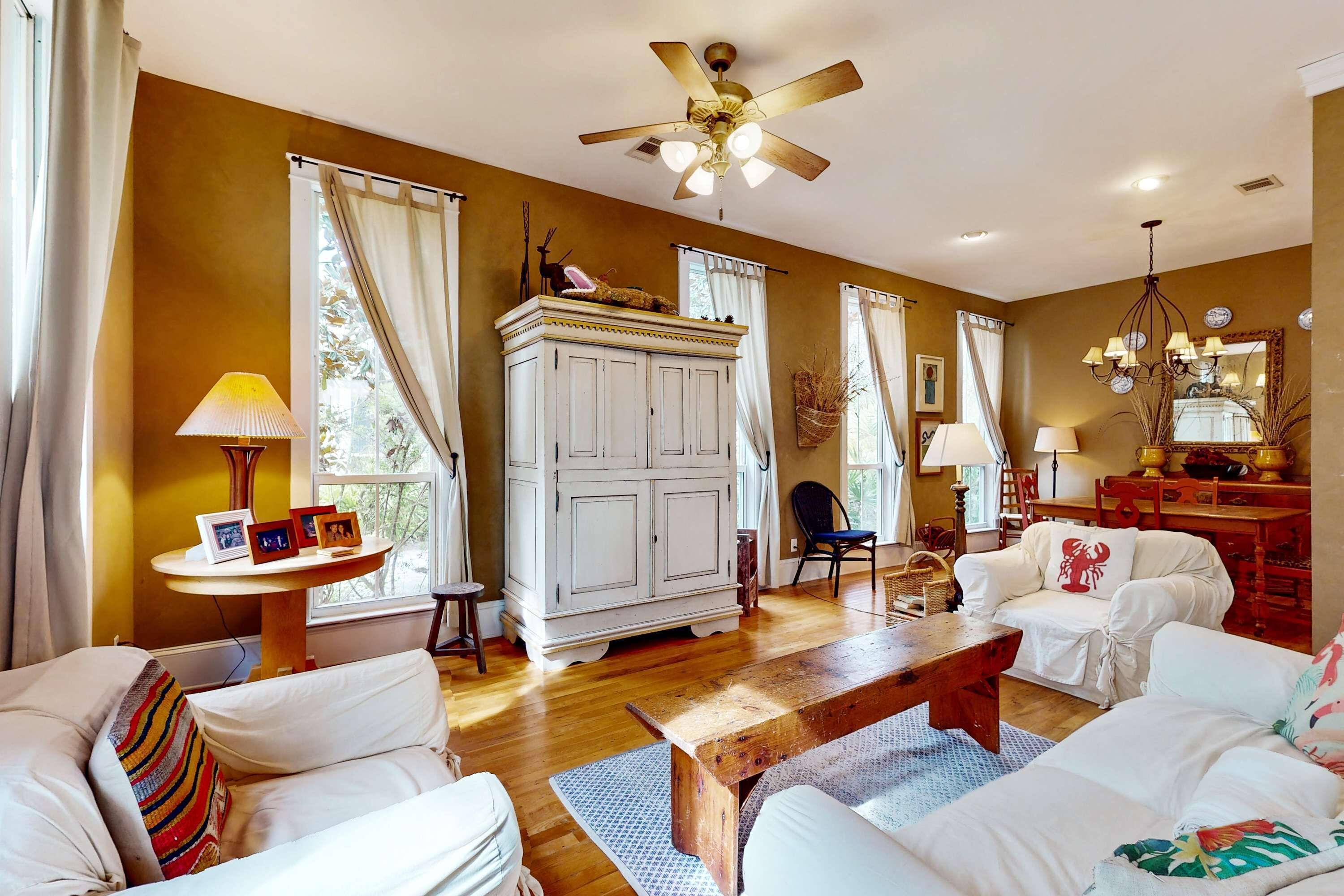 A spacious living room with a wooden floor, white furniture, a ceiling fan, and large windows with curtains.