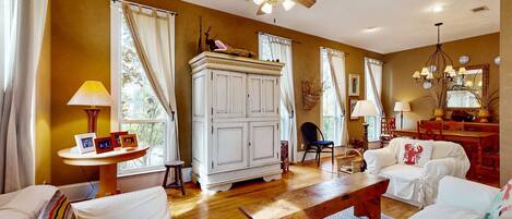 A spacious living room with a wooden floor, white furniture, a ceiling fan, and large windows with curtains.