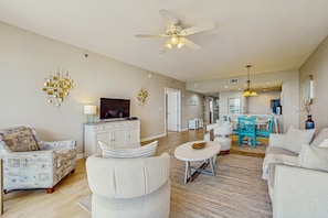 A spacious living room with a ceiling fan, a television, and a dining area with a table and chairs. - High rise beach condo w water views & swimming pool - close to local sights (Destin)