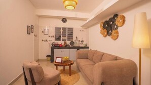Un espacio habitable compacto con sofá, sillón y una pequeña mesa redonda. Se observa una cocina al fondo, con una ventana y un salpicadero de azulejos blancos.