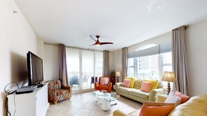 Un salón amplio con una televisión de pantalla plana, un ventilador de techo y un ventanal con cortinas.