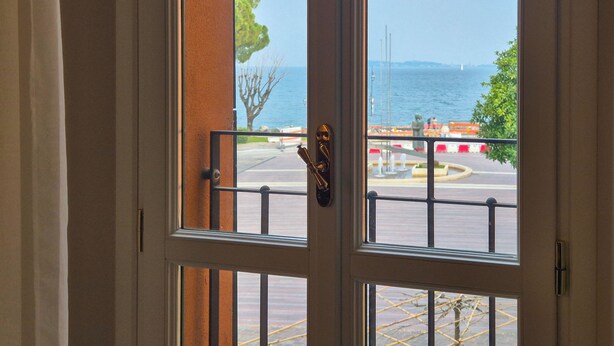 Blick durch ein Fenster mit Metallgitterstäben, einer Tür und einem Blick auf ein Gewässer sowie eine Parkanlage.