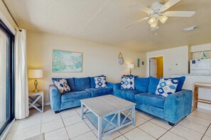 A living room with a blue sofa set, a wooden coffee table, a ceiling fan, and a painting on the wall. - Gorgeous, Open Ground Floor Condo Gulf Front w Large Patio &  Beach Access (Destin)