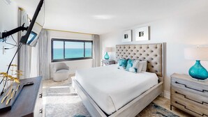 Ett hotellrum med en stor säng, ett skrivbord med en lampa, en stol, ett fönster med utsikt över havet och en taklampa.