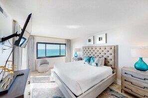 Une chambre d’hôtel avec un grand lit, un bureau avec une lampe, une chaise, une fenêtre donnant sur la mer et un luminaire fixé au plafond.