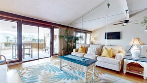 Ein geräumiges Wohnzimmer mit einer großen Schiebeglastür, die auf einen Balkon führt, ein weißes Sofa mit gelben Kissen, ein Couchtisch und ein Deckenventilator.