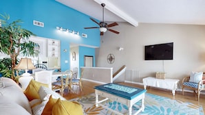 Ein Wohnzimmer mit blauer Akzentwand, einem Deckenventilator, einem Fernseher, einem Couchtisch und einer Treppe, die zu einer weiteren Ebene führt.