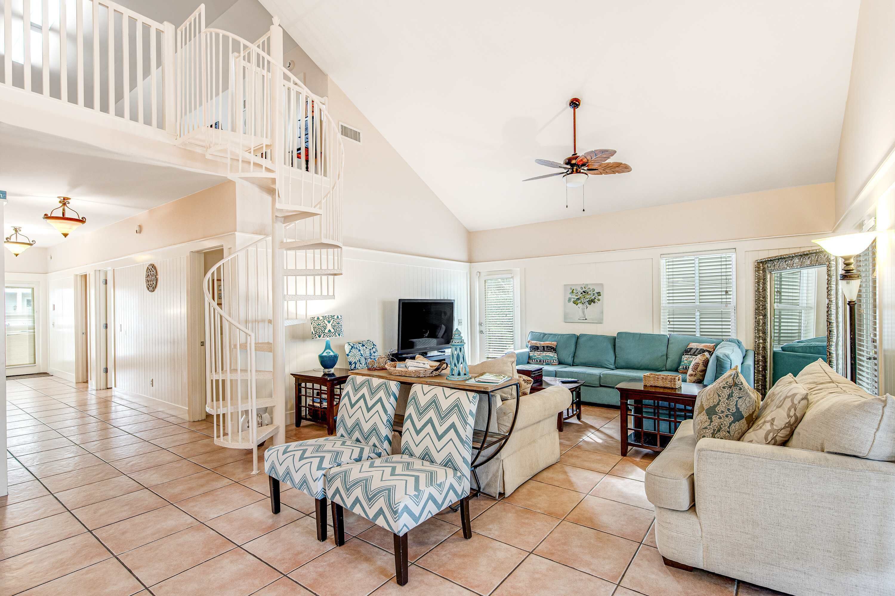 A spacious living room with a white staircase, a ceiling fan, and a teal sofa.