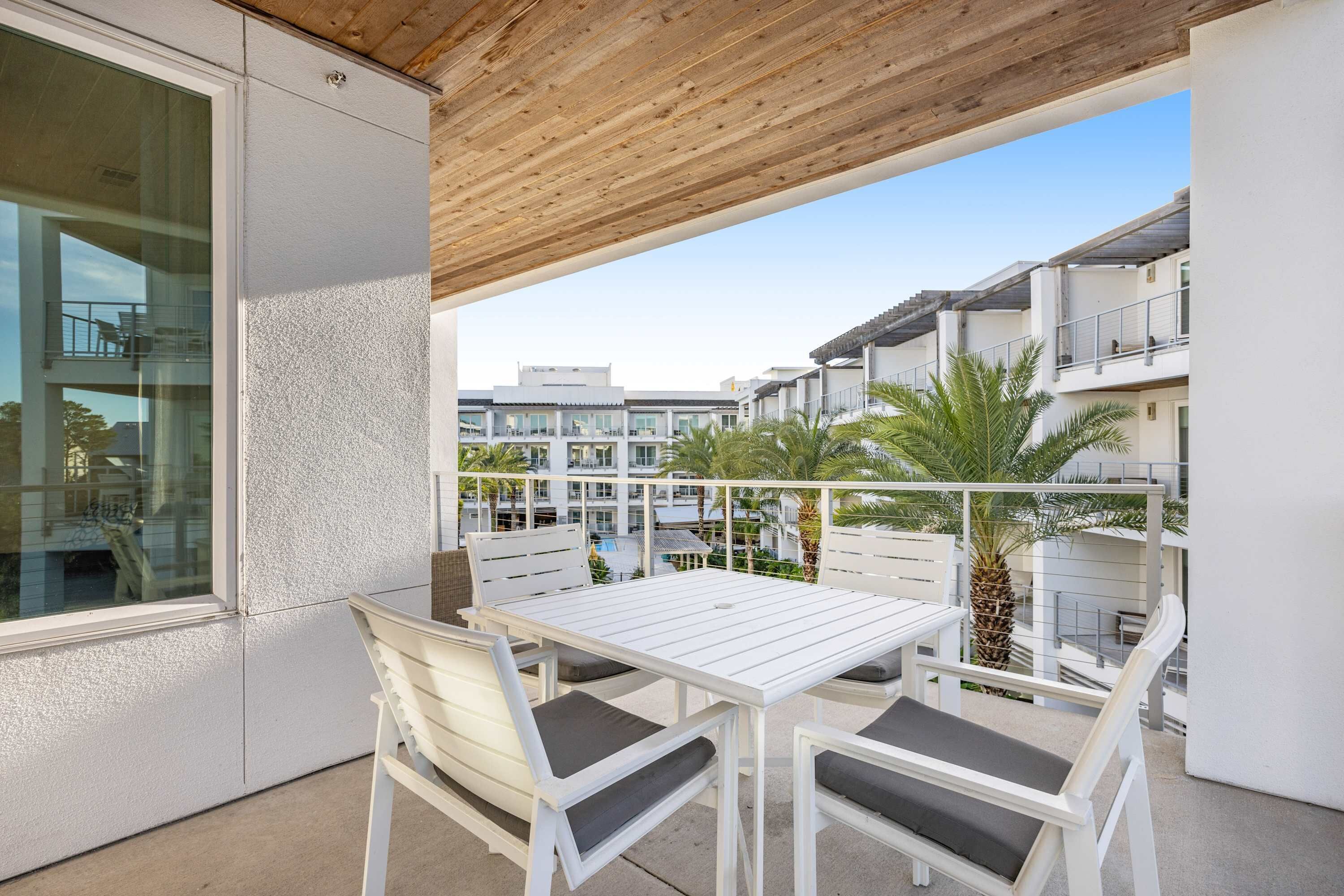 A balcony with a white table and chairs, overlooking a residential area with palm trees and modern buildings.