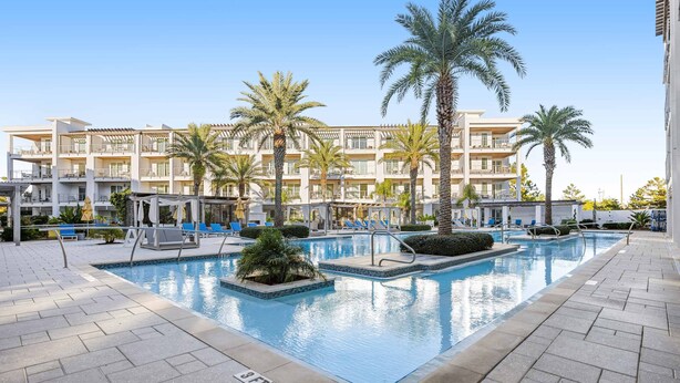Una piscina con sillones de descanso y palmeras, rodeada por un edificio moderno con balcones.