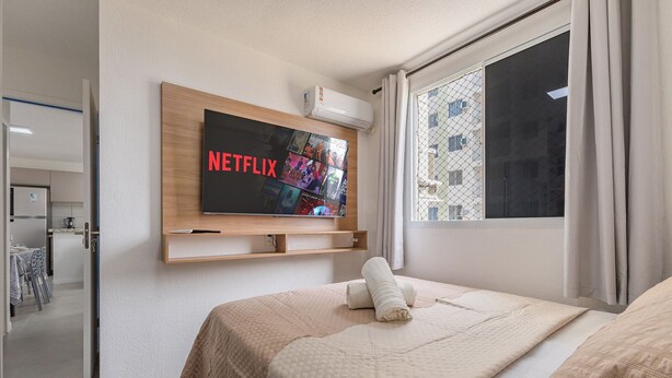 A hotel room with a large bed, a mounted TV displaying the Netflix logo, a window with curtains, and a view of a building outside. - Mirante #801 - Two Bedroom Apt by Carpediem (Fortaleza)