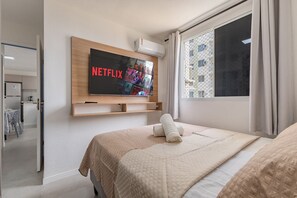 Habitación de hotel con una cama grande, un televisor montado mostrando el logotipo de Netflix, una ventana con cortinas y vistas a un edificio.