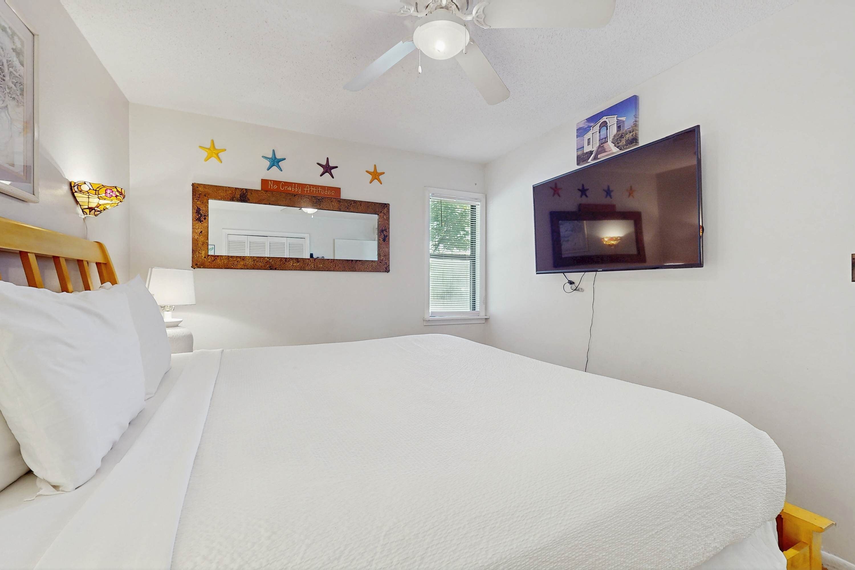 A bedroom with a bed, a ceiling fan, a TV, a window with blinds, and a wall with star decorations and a mirror.