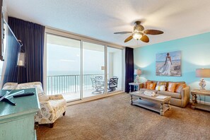 Ein geräumiges Wohnzimmer mit einer großen Glasschiebetür, die auf einen Balkon führt, einem Deckenventilator, einem Sofa, einem Couchtisch und einem Bild an der Wand.