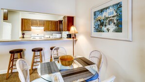 Una cucina compatta con mobili in legno, un frigorifero bianco e un forno a microonde. C'è una zona pranzo con un tavolo in vetro, sedie bianche e una ciotola di legno. Un quadro appeso è sulla parete.