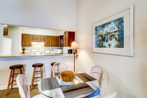 Une cuisine compacte avec des meubles en bois, un réfrigérateur blanc et un four à micro-ondes. On y trouve un coin repas avec une table en verre, des chaises blanches et un saladier en bois. Un tableau encadré est accroché au mur.