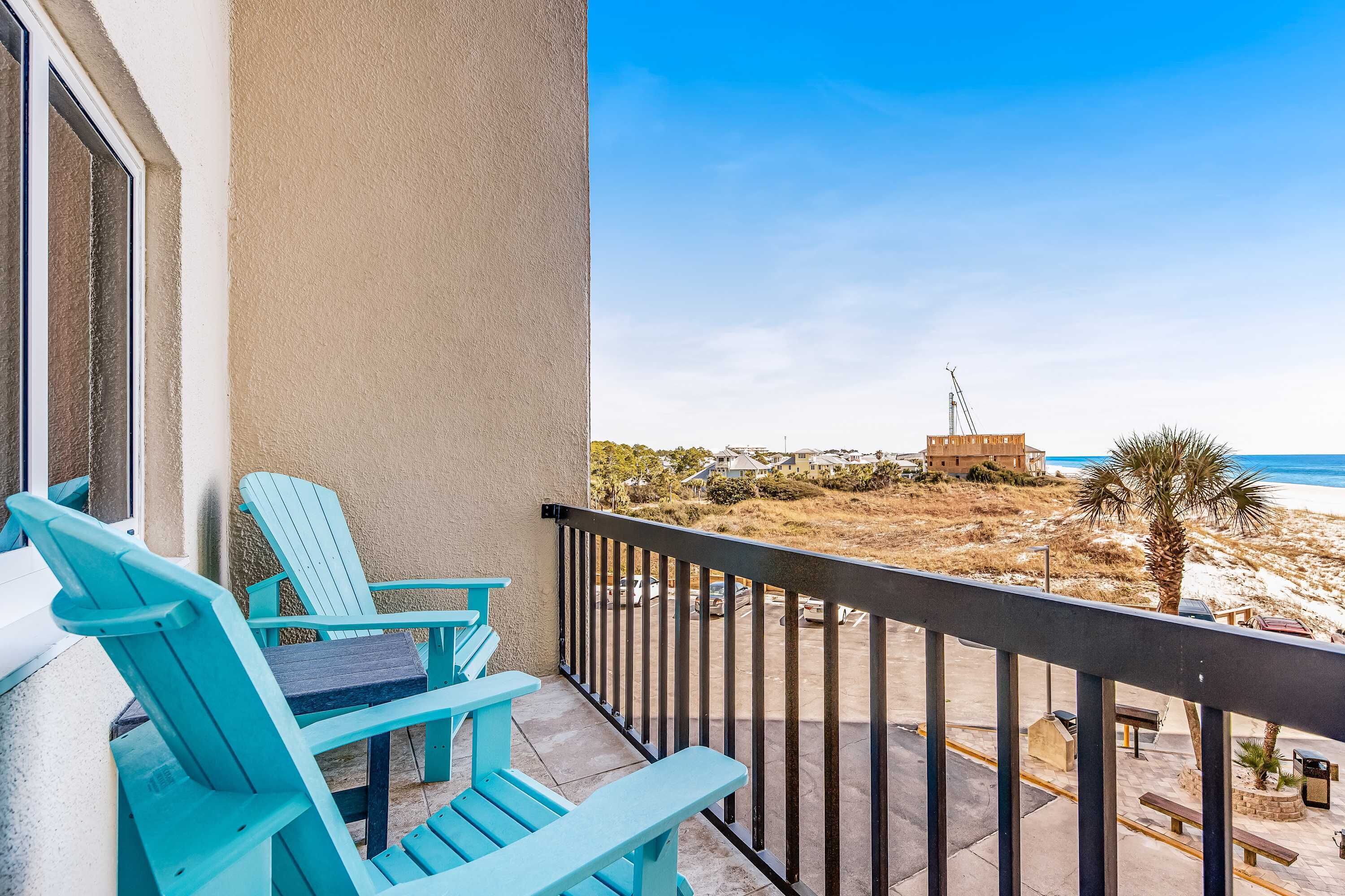 Un balcon avec des fauteuils Adirondack turquoise, une vue sur la plage et un bâtiment situé au loin.