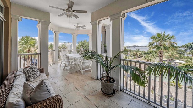 A balcony with a ceiling fan, a potted plant, a sofa with cushions, and a view of palm trees and a body of water. - Luxurious bayfront condo in a gated resort with an amazing pool & hot tub (Panama City Beach)