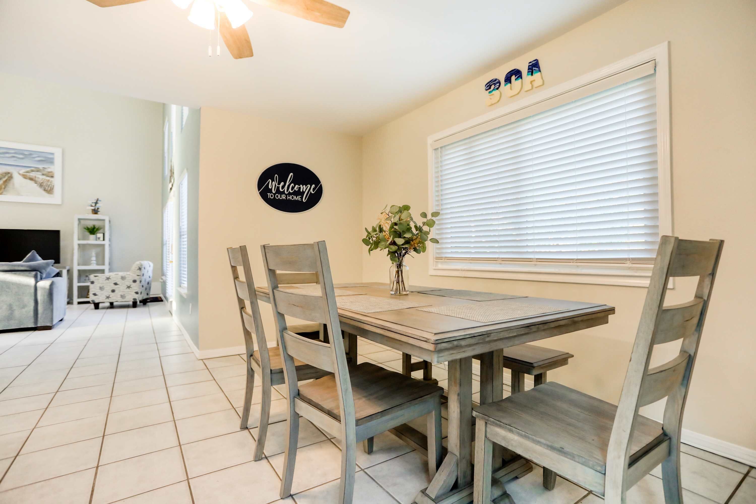 A dining area with a wooden table and chairs, a vase with flowers, and a 'Welcome to our home' sign on the wall.