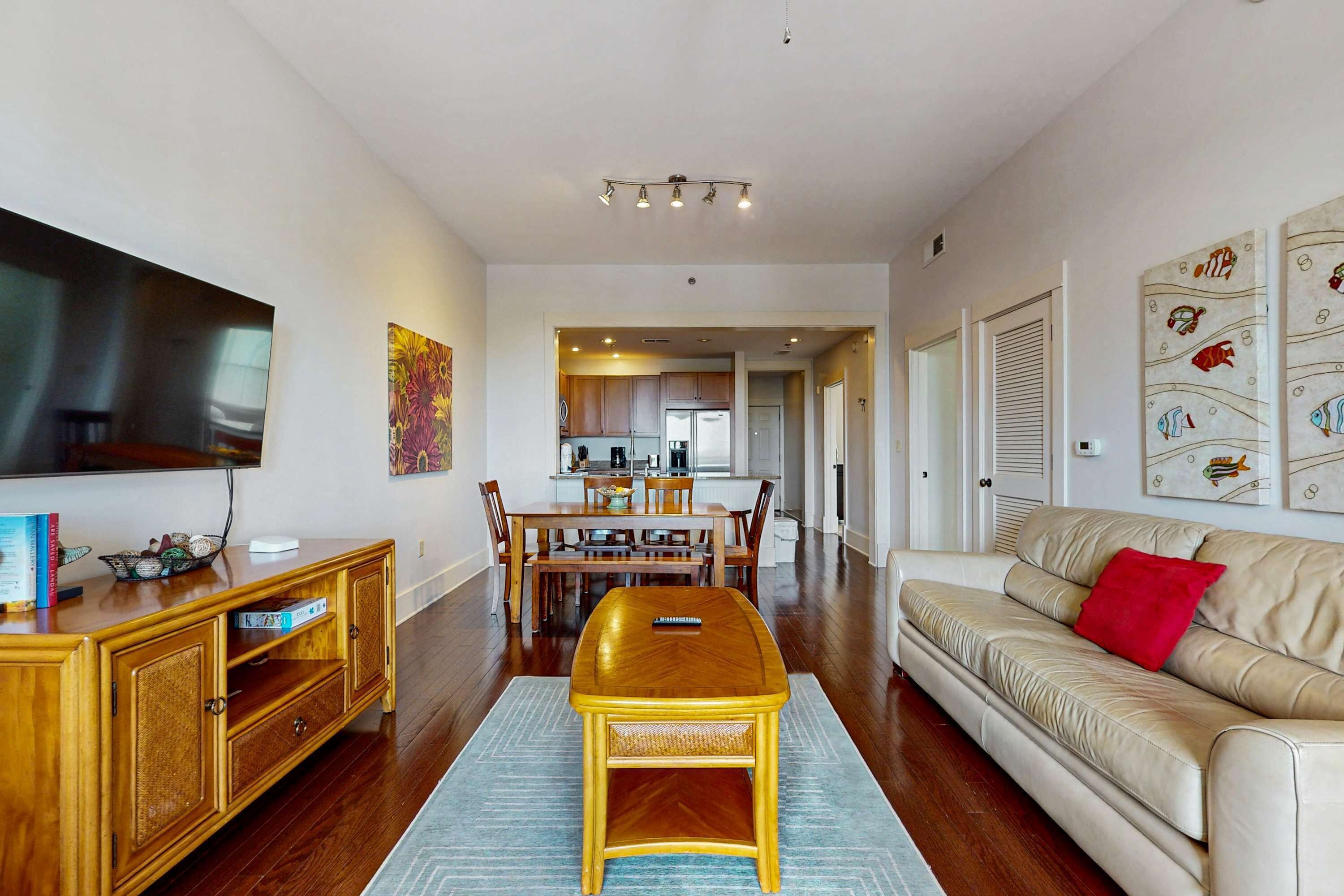 A living room with a wooden entertainment center, a beige sofa with a red pillow, a dining area with wooden furniture, and a kitchen in the background.