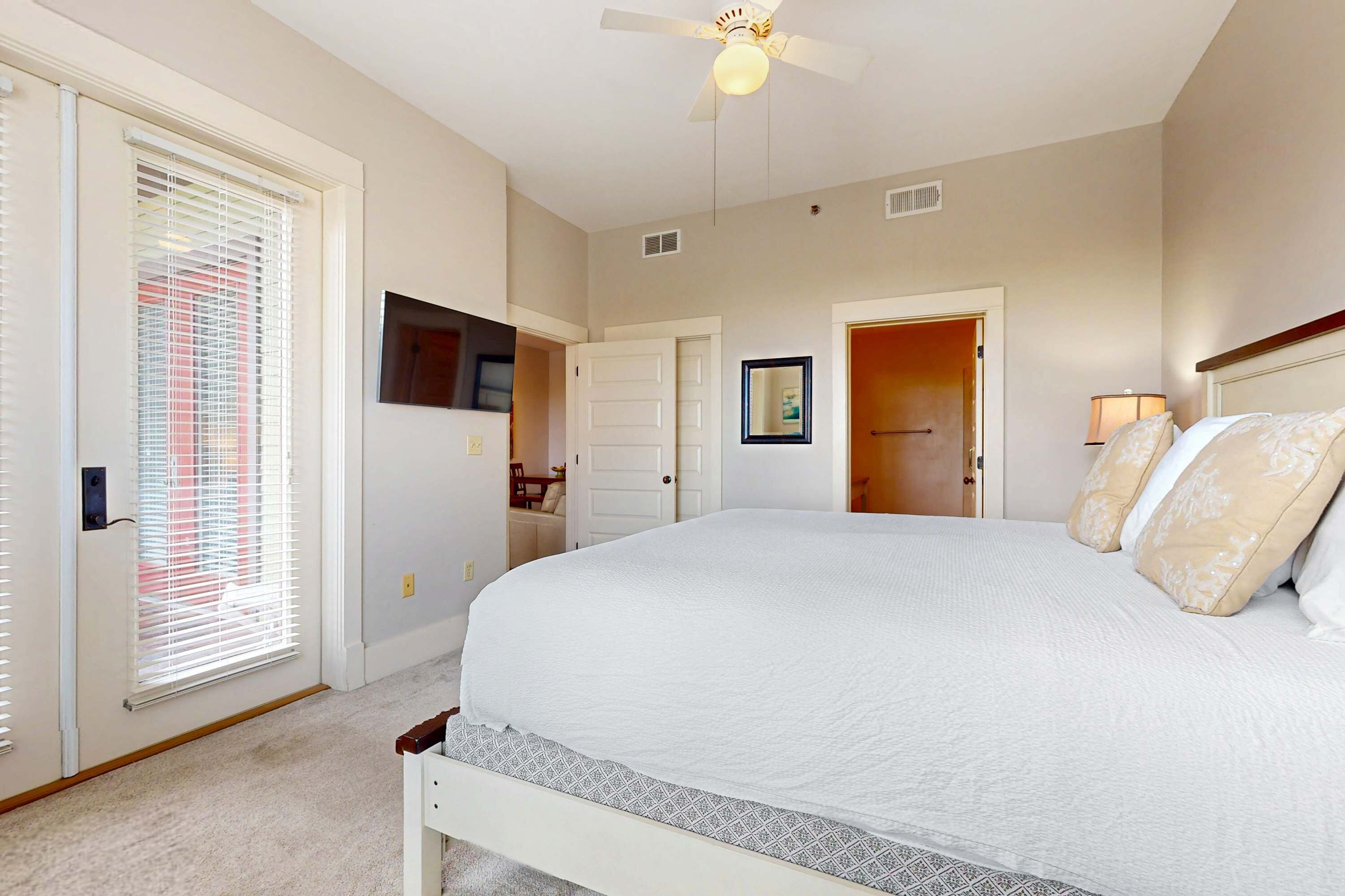 A bedroom with a large bed, a ceiling fan, a TV mounted on the wall, and a door leading to another room.