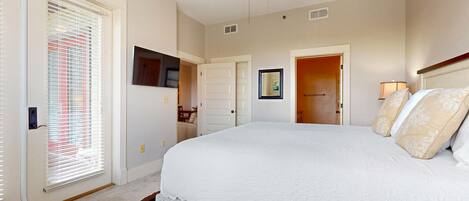 A bedroom with a large bed, a ceiling fan, a TV mounted on the wall, and a door leading to another room.