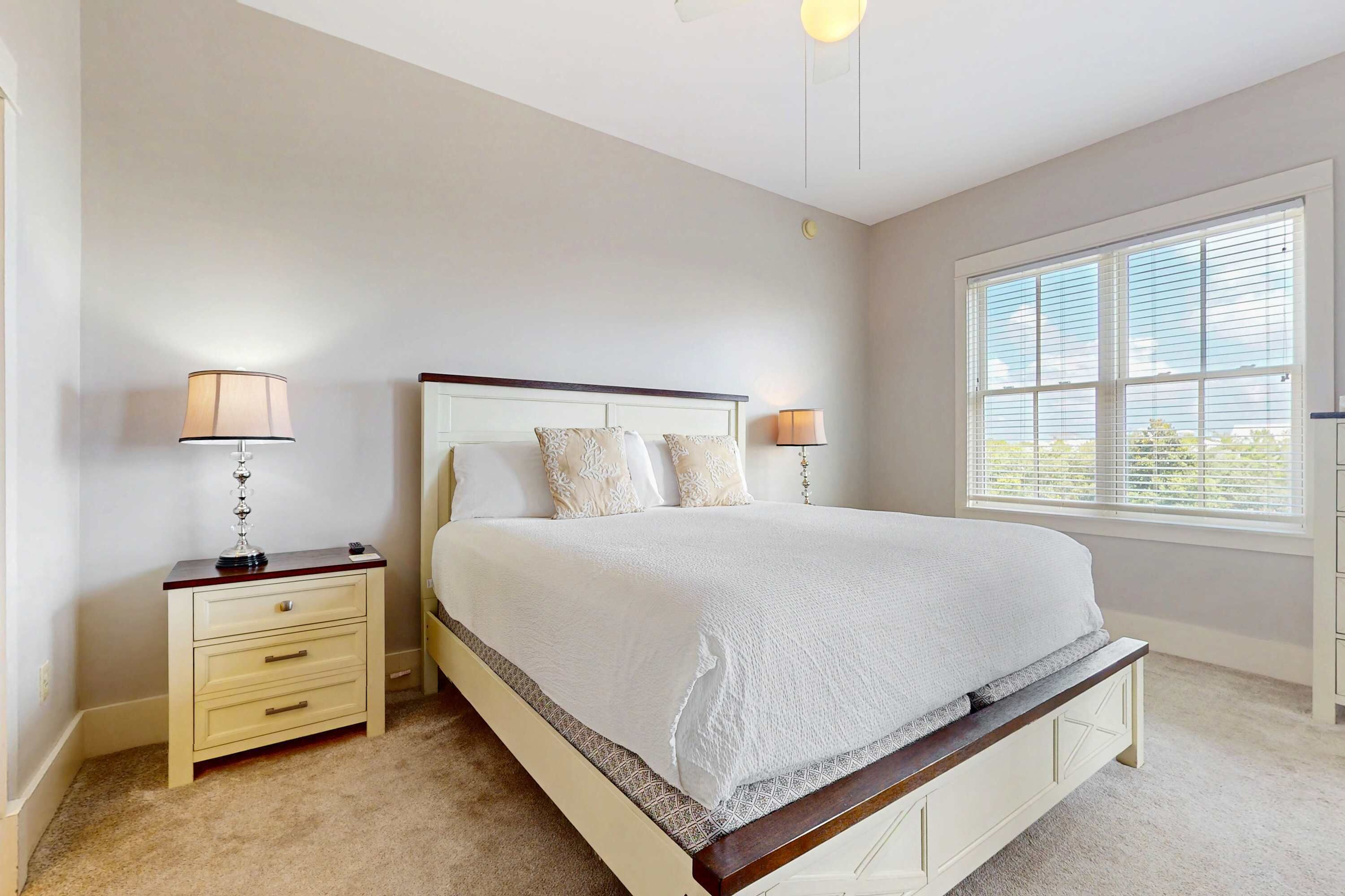 A bedroom with a large bed, two bedside tables with lamps, a window with blinds, and a carpeted floor.