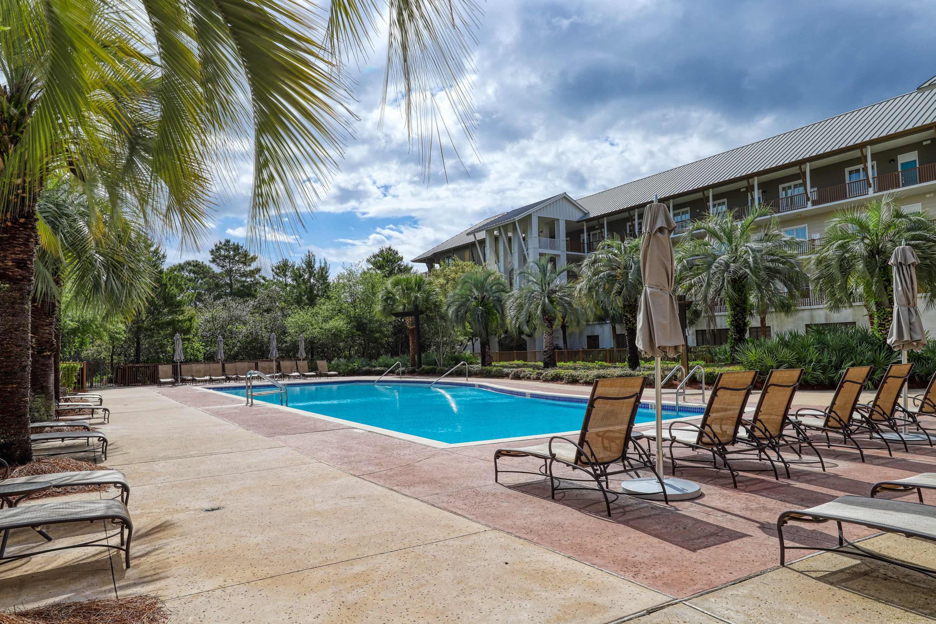 A swimming pool with lounge chairs and umbrellas, surrounded by palm trees and a building with balconies.