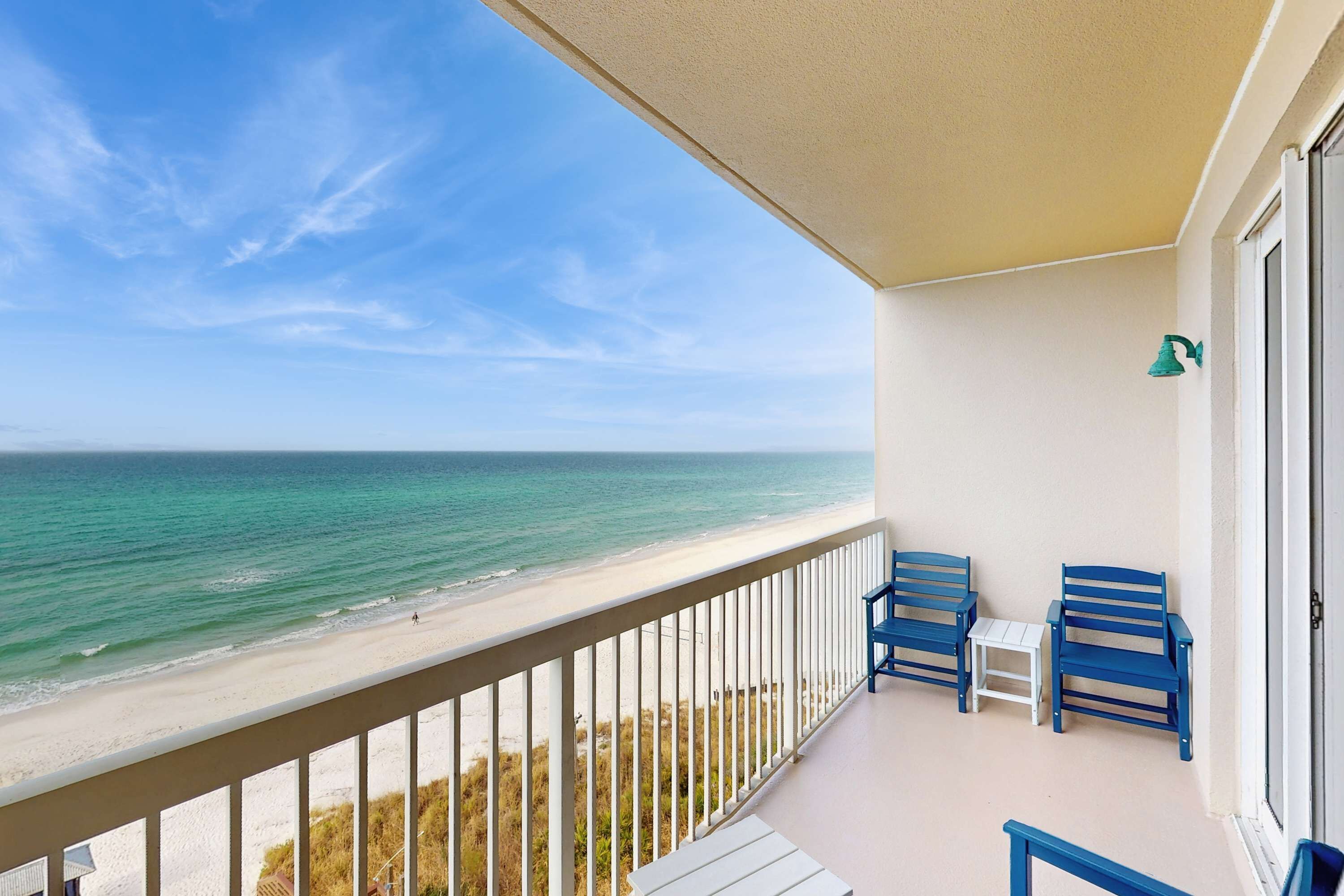 A balcony with a view of the beach and ocean, two blue chairs, and a small table.