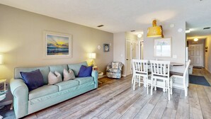 A living room with a light blue sofa, white dining table, and chairs. There are wall-mounted lamps, a framed picture, and a ceiling fan. - Lovely second-floor condo w/beach access & shared outdoor pool (Panama City Beach)