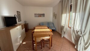 A room with a wooden dining table, chairs, a TV on a cabinet, a bed with a blue blanket, and a window with curtains. - Cozy Flat With Private Parking in Rosolina Mare (Rosolina)