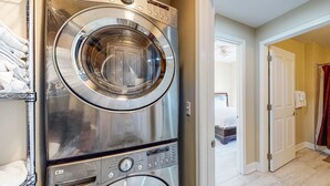 A stacked washer and dryer unit in a laundry room with a bed visible in the background. - Snowbird-friendly condo with waterfront view, WiFi, AC, access to pool, hot tub (Panama City Beach)