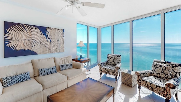 A spacious living room with a large window overlooking the ocean, a ceiling fan, and a modern sofa with patterned cushions. - Bright Gulf-facing condo on the beach with pool & hot tub (Panama City Beach)