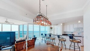 A spacious dining area with a large wooden table and chairs, a hanging decorative light fixture, and a view of the ocean through large windows. - Bright Gulf-facing condo on the beach with pool & hot tub (Panama City Beach)