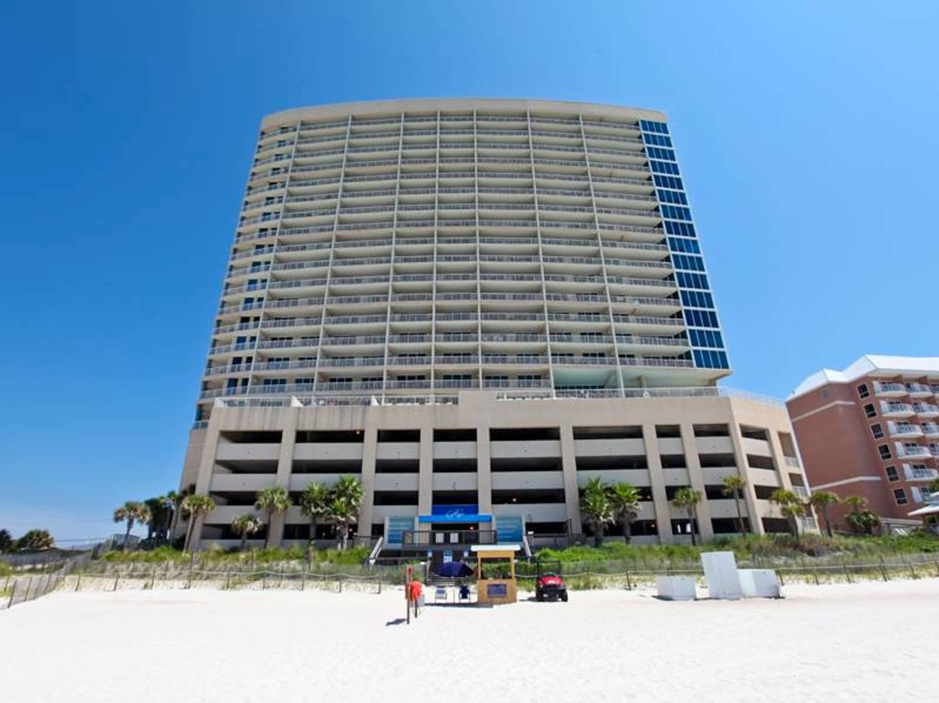 A tall, multi-story hotel with a curved facade, a parking garage below, and a beachside location with palm trees.