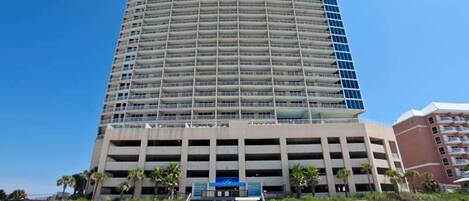 A tall, multi-story hotel with a curved facade, a parking garage below, and a beachside location with palm trees.