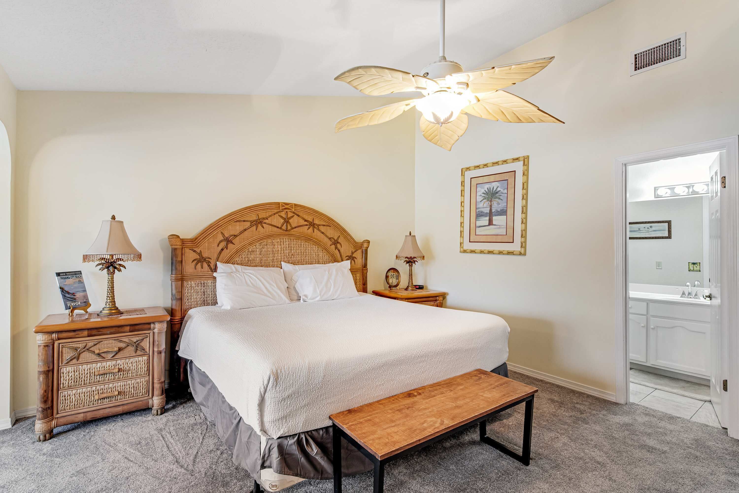 A bedroom with a large bed, wooden headboard, two bedside tables with lamps, a bench, a ceiling fan, and a bathroom visible through an open door.