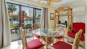 A room with a glass-top table surrounded by wicker chairs with red cushions, a ceiling fan, and a view of a pool and palm trees through a sliding glass door. - Conveniently located condo with shared pool & tennis, on-site golf - walk to beach (Panama City Beach)