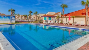 Una piscina con lettini da relax e palme, circondata da edifici con tetti in tegole rosse.