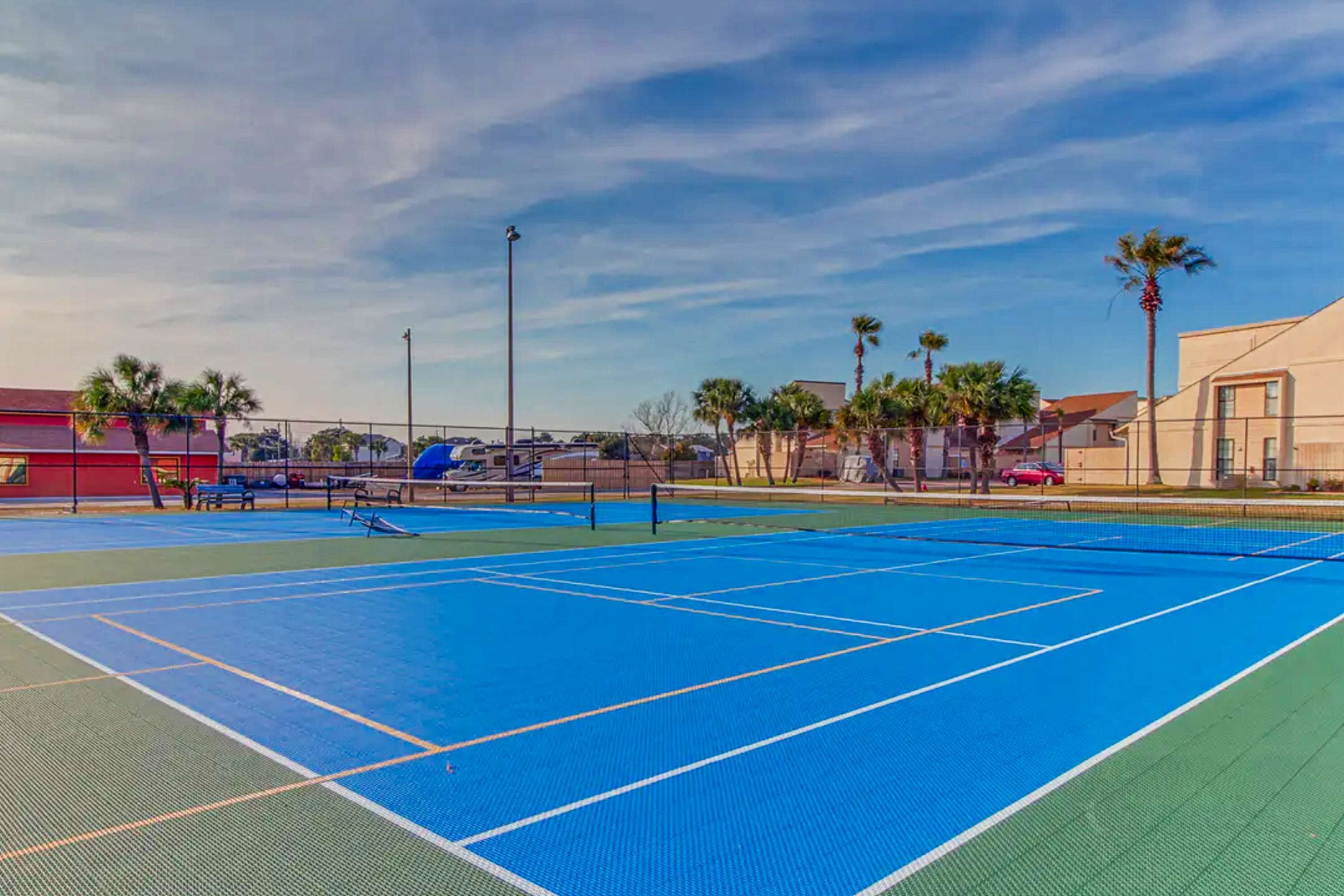 Ein Tennisplatz im Freien mit blauer Belag, umgeben von Palmen und Gebäuden.