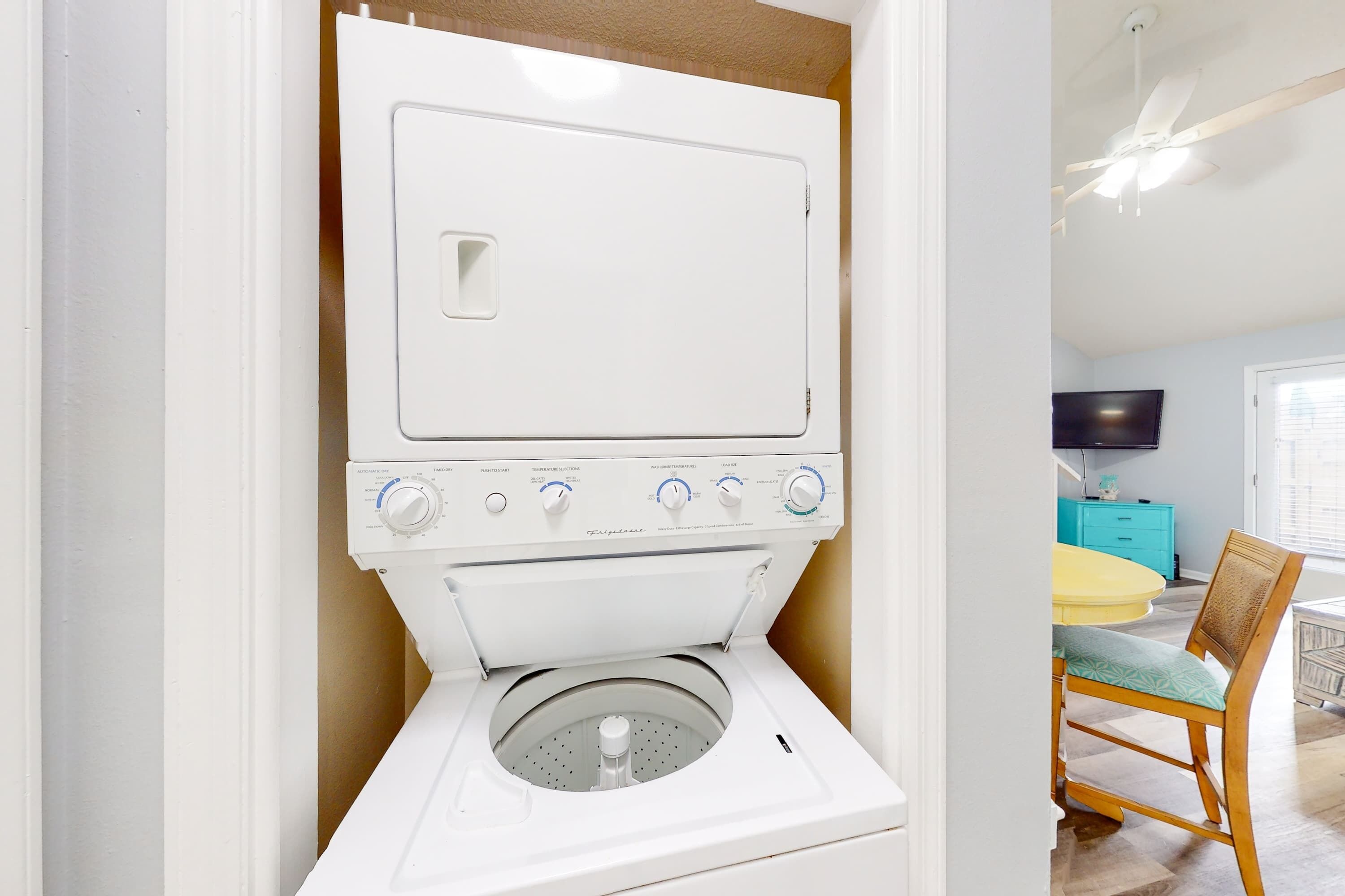 Eine kompakte, weiße Waschmaschine mit Frontlader in einem gut beleuchteten Raum, mit einem Holzesstisch und -stühlen, einem Flachbildfernseher an der Wand und einem Deckenventilator.