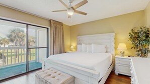 Een slaapkamer met een groot bed, een plafondventilator, een nachtkastje met lamp, een kamerplant en een glazen schuifdeur naar een balkon.