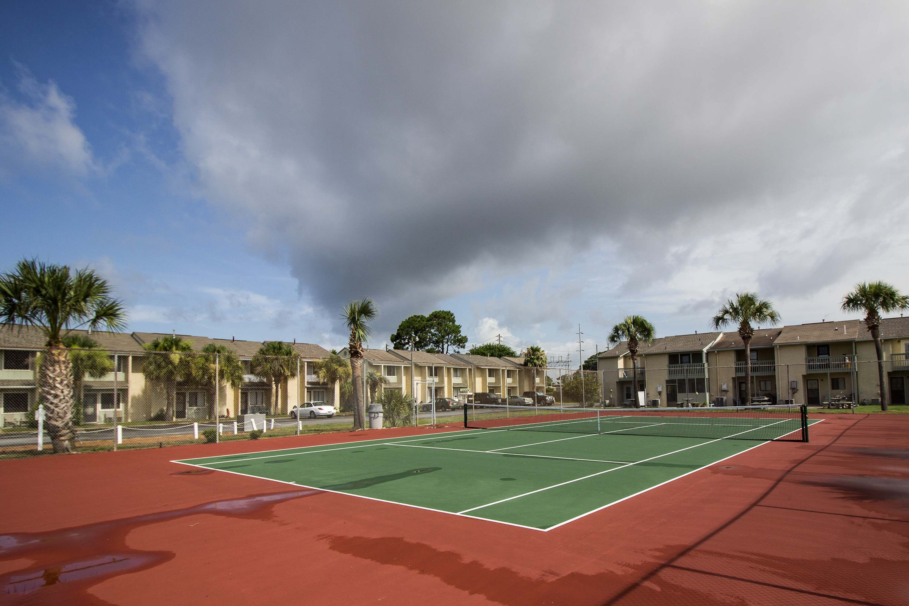 An outdoor tennis court with a green playing surface and red surrounding area, surrounded by buildings and palm trees.
