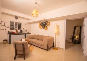Un salon avec un canapé beige, une chaise en bois, une table basse ronde, un lampadaire et un coin cuisine comprenant une fenêtre, un évier et des placards.