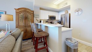 Una cocina compacta con armarios blancos, un microondas y un refrigerador. Hay un mueble de madera con una silla tejida al frente. Un bote de basura es visible cerca del mostrador de la cocina.