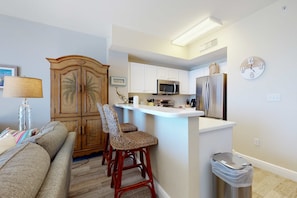 A compact kitchen with white cabinets, a microwave, and a refrigerator. There is a wooden cabinet with a woven chair in front. A trash bin is visible near the kitchen counter. - Oceanfront condo with scenic balcony & washer/dryer - pool/hot tub/tennis access (Panama City Beach)