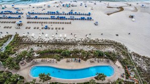 Vista aérea de una playa con filas de tumbonas y sombrillas, una piscina rodeada de palmeras, y una zona de arena con gente y sillas de playa.