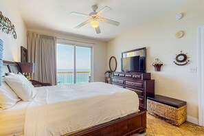 A hotel room with a bed, a dresser, a television, a ceiling fan, a window with curtains, and a view of the ocean. - Gulf-view condo with pools & balcony - walk to Pier Park (Panama City Beach)